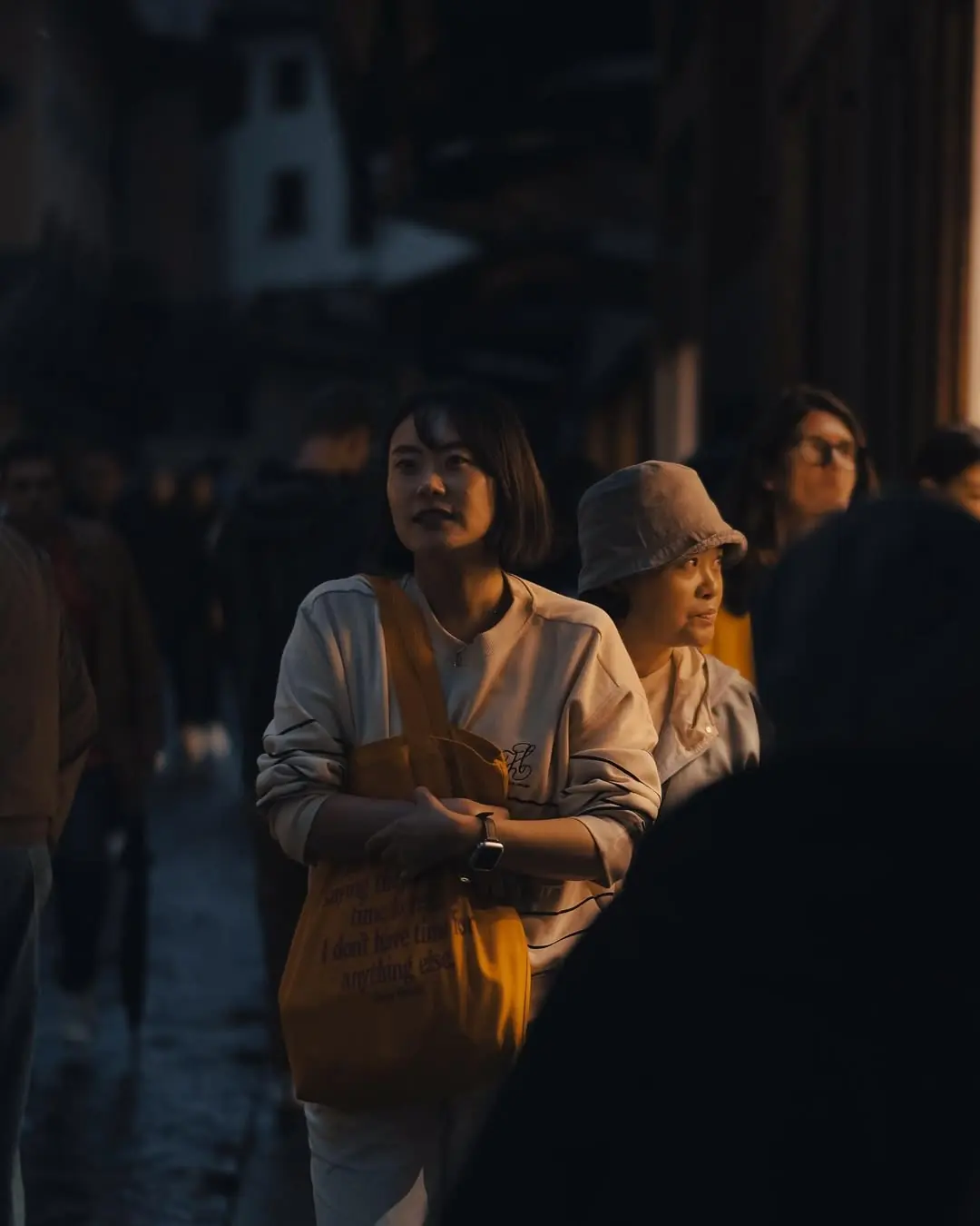 Two women pause in a crowded street at golden hour, side-lit, a yellow tote bag catching the last of the light.