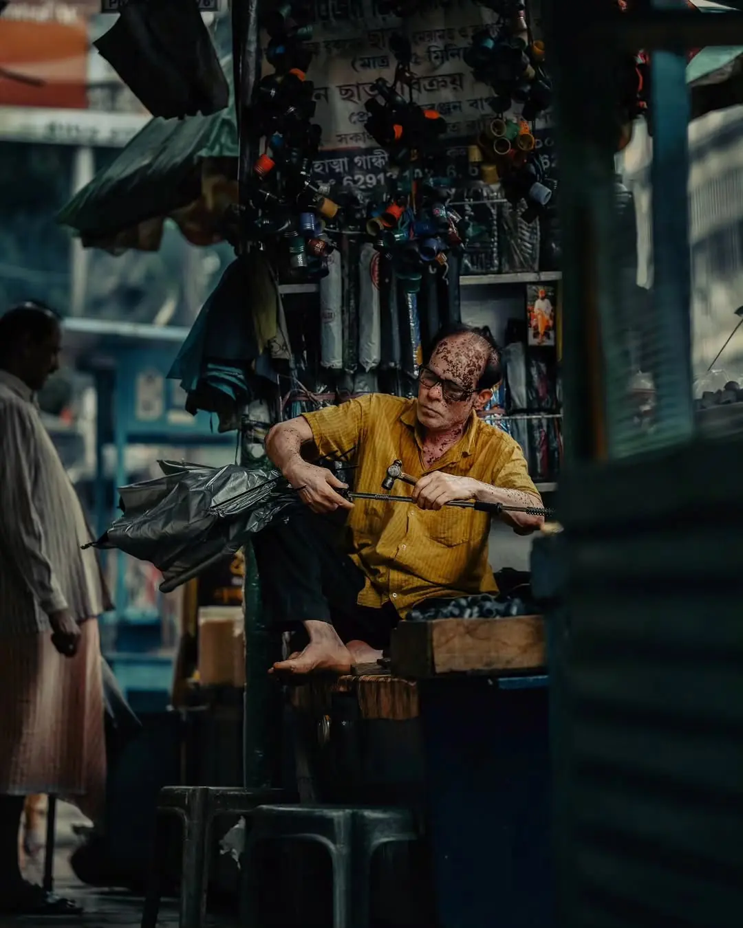 An umbrella repairer works barefoot at his roadside stall, tapping a spoke into place beneath a canopy of hanging handles.