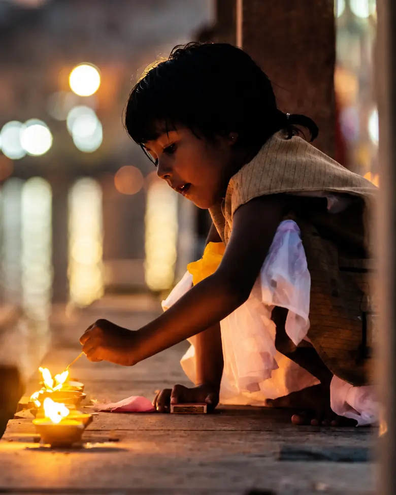 A small girl crouches on a ghat at dusk lighting oil lamps, the lights of the far bank blurred behind her.
