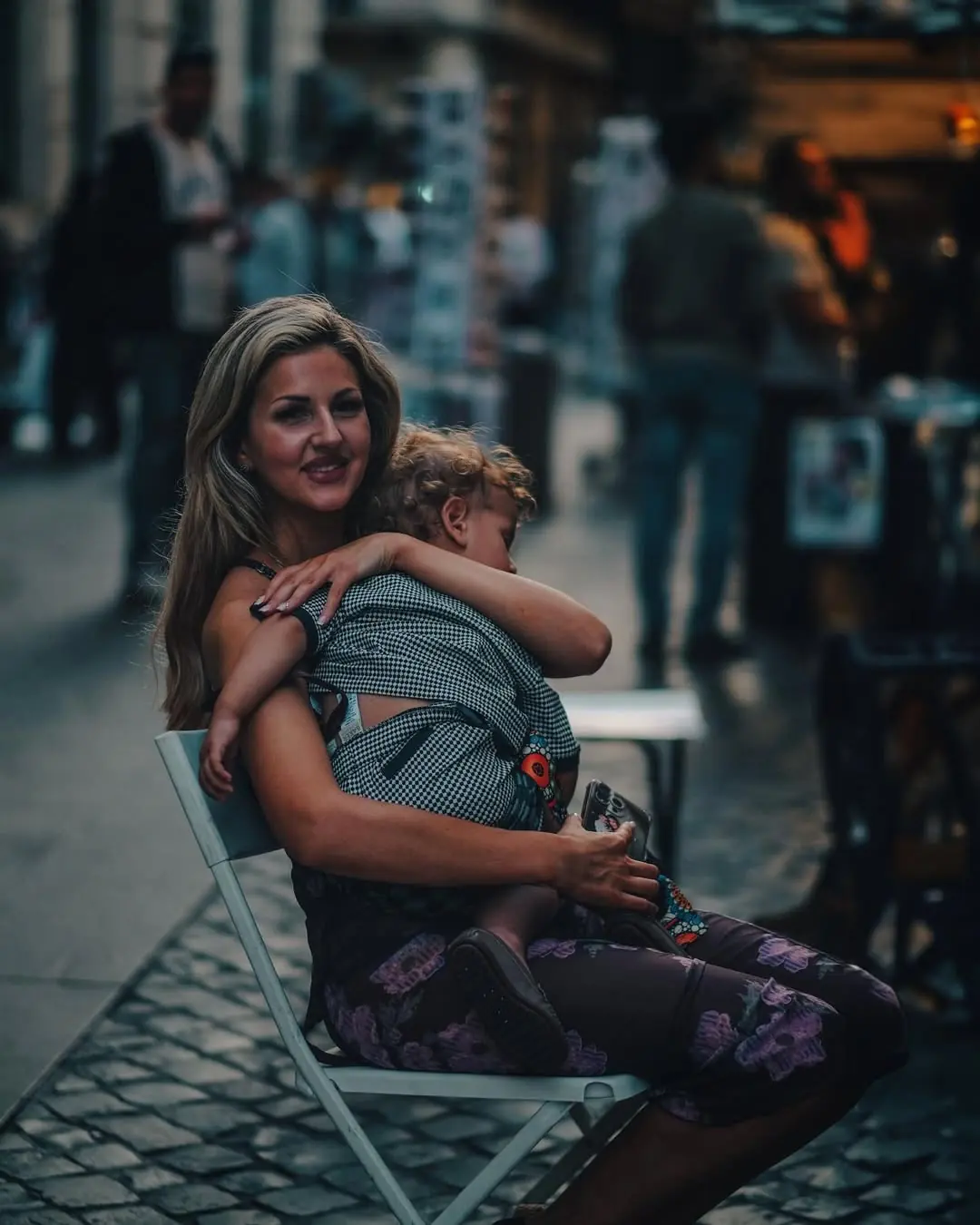 A woman sits in a folding chair on a cobbled street holding a sleeping toddler, smiling past the camera.